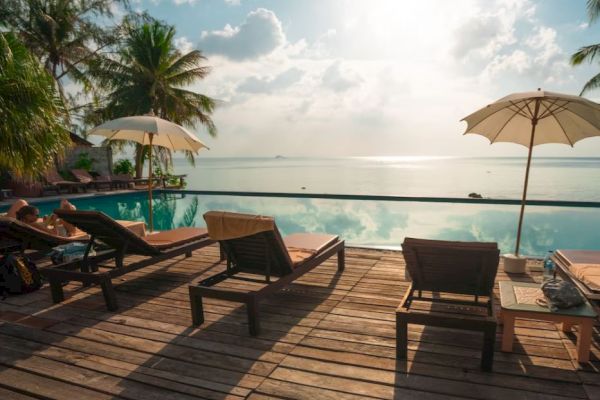 A serene poolside scene with lounge chairs, umbrellas, palm trees, and a view of the ocean under a bright, partly cloudy sky.