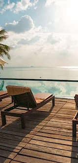 Poolside scene with lounge chairs, umbrellas, and palm trees overlooking a serene body of water under a partly cloudy sky.