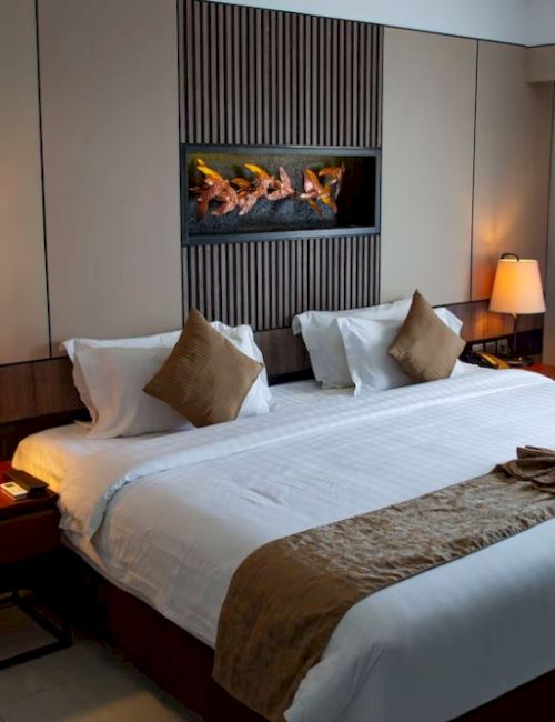 A neatly arranged hotel room with a large bed, two bedside lamps, and a fireplace artwork above the bed. Curtains are drawn to the side.