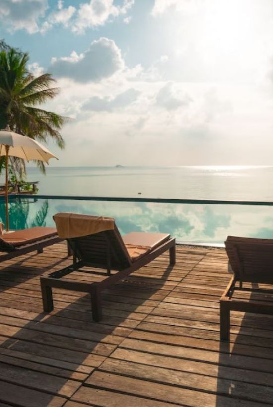 This image shows a serene poolside with lounge chairs, umbrellas, and palm trees overlooking a calm body of water under a partly cloudy sky.