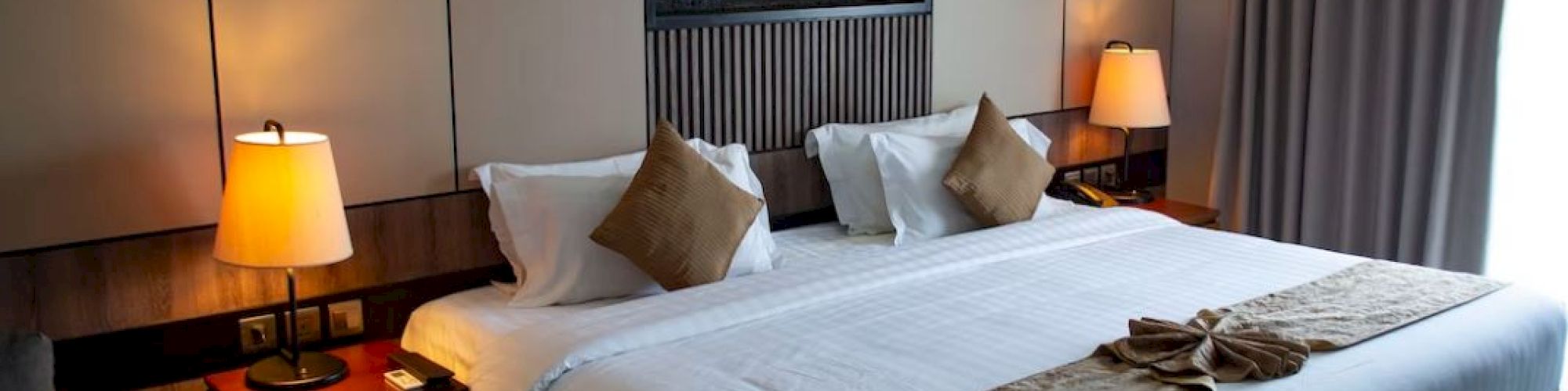 A cozy hotel room with a large bed, pillows, lamps, and a decorative panel above the headboard. A towel arrangement on the bed.