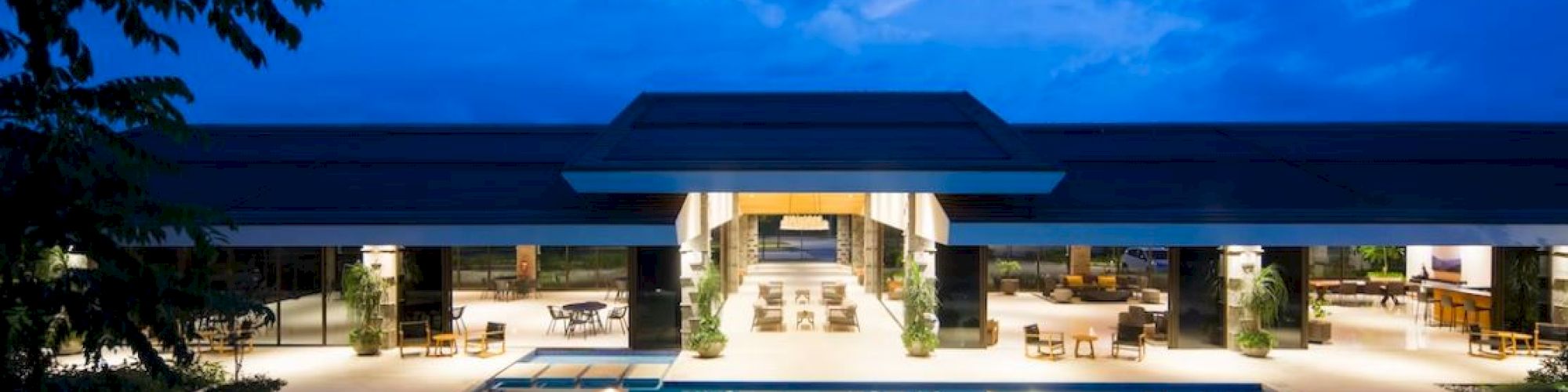 The image shows a luxurious modern building with a large illuminated swimming pool in the foreground during twilight.