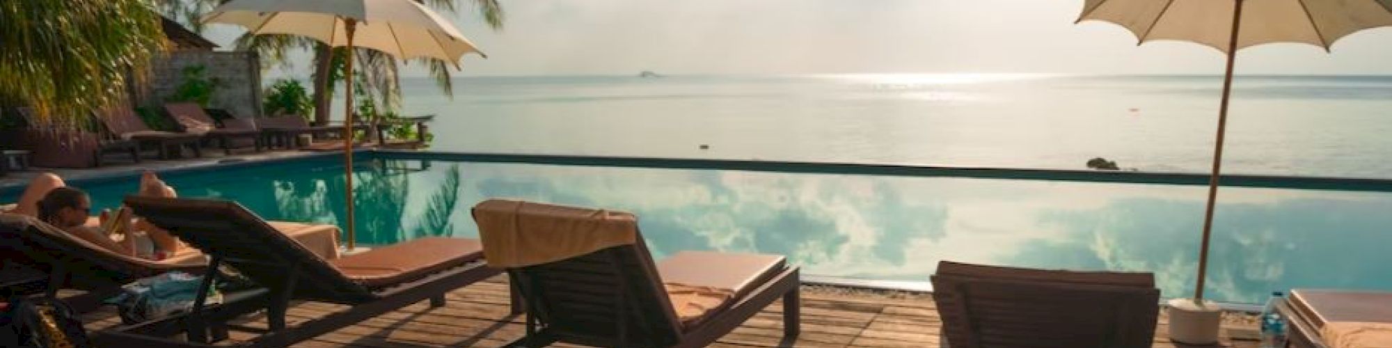 A serene poolside scene with lounge chairs, umbrellas, palm trees, and a tranquil ocean view under a partly cloudy sky and sunlight.