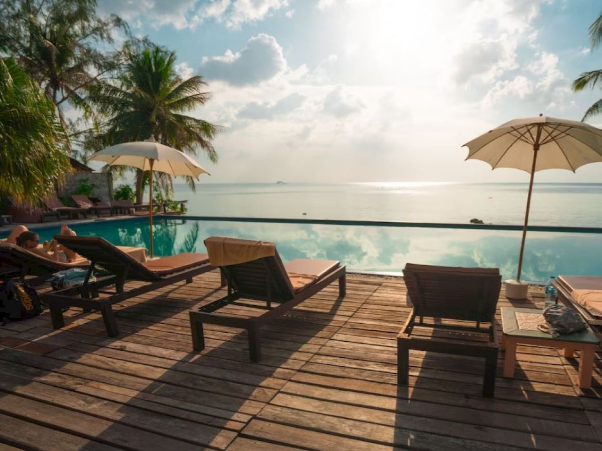 A serene beach scene with lounge chairs, umbrellas, a wooden deck, and a pool overlooking the ocean under a bright, cloudy sky.