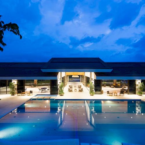 A luxurious illuminated pool extends before a modern open-air structure under a twilight sky.