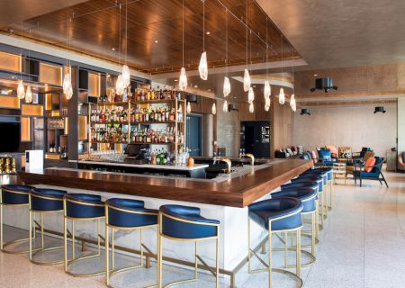 The image shows a modern bar with overhead lights, a stocked back shelf, blue bar stools, and a lounge area with additional seating.