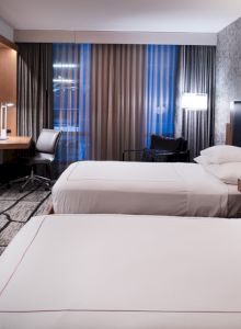 A modern hotel room with two beds, a desk, a chair, a TV on the wall, and stylish decor with patterned wallpaper and carpet.