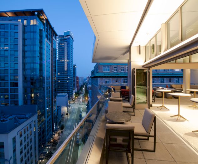 A modern cityscape at dusk with high-rise buildings and a stylish outdoor balcony featuring seating and tables.