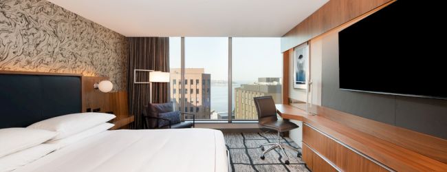 The image shows a modern hotel room with a large bed, desk, TV, and a window with a city view.