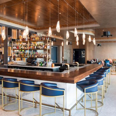 A modern bar with stylish blue stools, pendant lights, a stocked back bar, and cozy seating in the background for a relaxing ambiance.