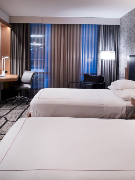 The image shows a modern hotel room with two beds, a wall-mounted TV, a desk with a chair, and stylish lighting.