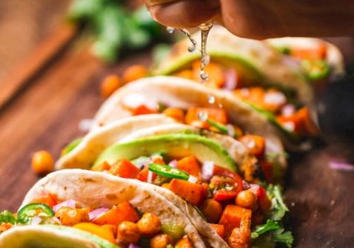 Tacos filled with chickpeas, diced vegetables, and garnished with avocado slices and herbs, being squeezed with lime.