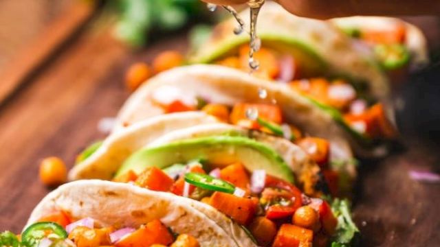 Tacos filled with chickpeas, diced vegetables, and garnished with avocado slices and herbs, being squeezed with lime.