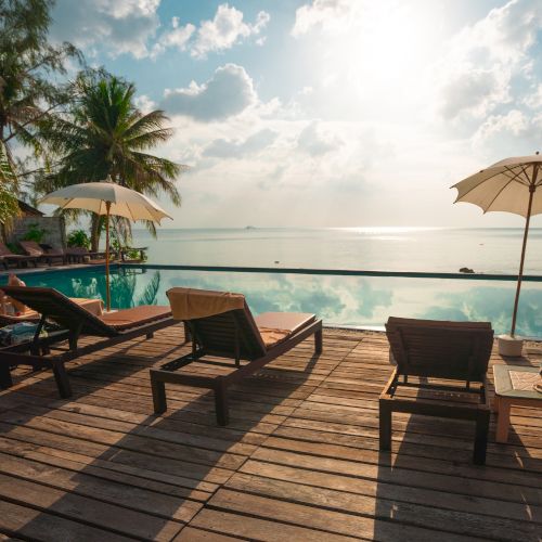 Lounge chairs and umbrellas by a poolside overlooking a serene ocean view, framed by palm trees under a bright sunlit sky.