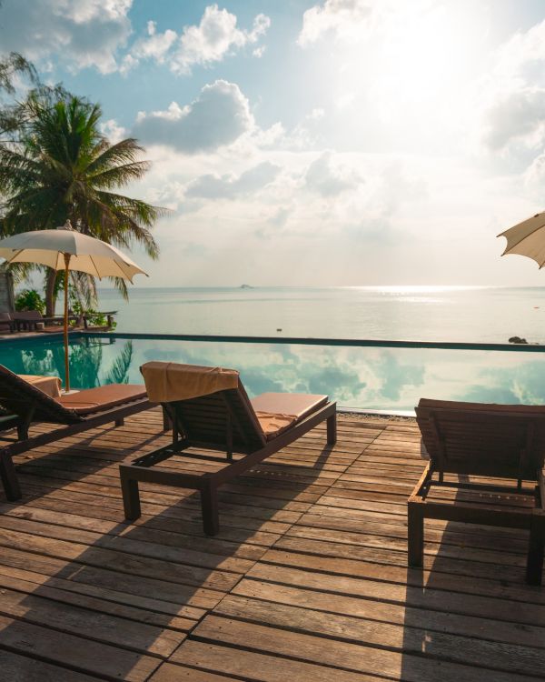 Lounge chairs and umbrellas by a poolside overlooking a serene ocean view, framed by palm trees under a bright sunlit sky.