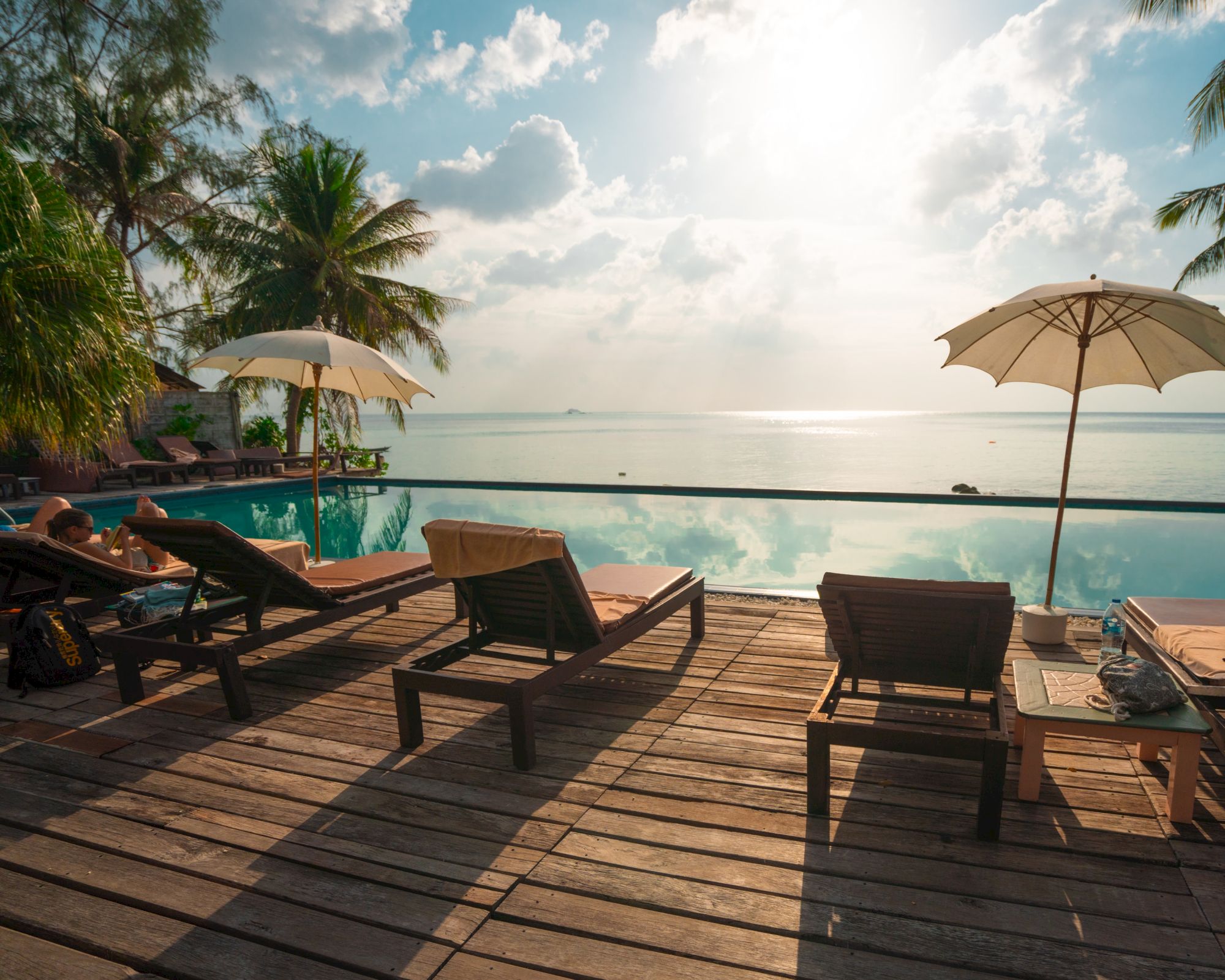 Sunlit beach scene with lounge chairs, umbrellas, palm trees, and an infinity pool overlooking a tranquil ocean view.