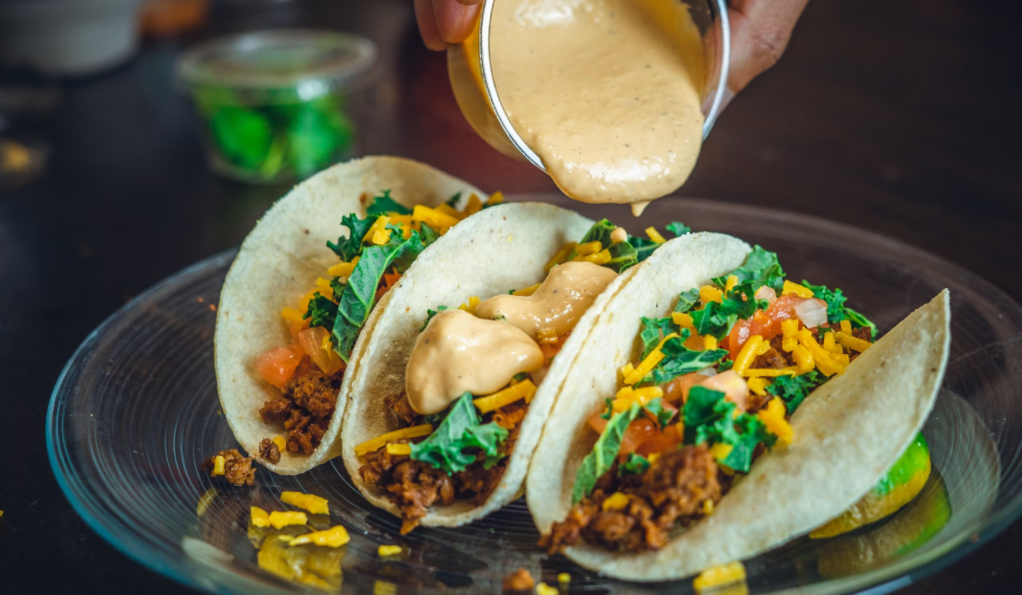Three tacos filled with meat, cheese, lettuce, and tomato are getting cheese sauce poured on them on a clear glass plate.
