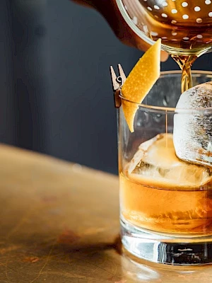 A drink being poured into a glass with ice and a citrus twist garnish on a bar counter.