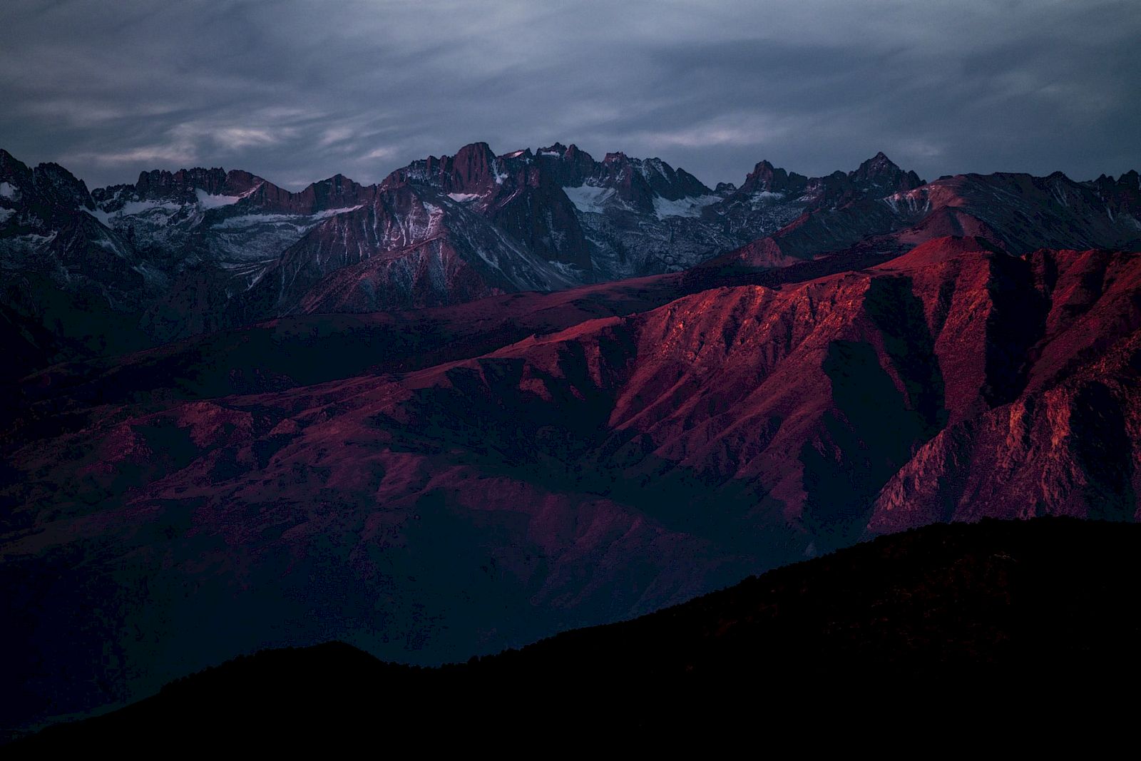 A mountain range is bathed in red light during sunset, with dramatic clouds above creating a striking contrast in the landscape.