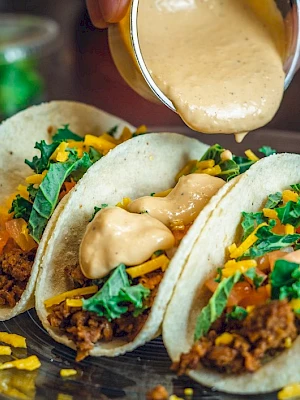 Three tacos with beef, cheese, tomatoes, and greens are being topped with a creamy sauce on a plate.