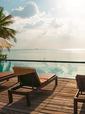 A serene poolside scene with lounge chairs, umbrellas, and a view of the ocean under a partly cloudy sky, surrounded by palm trees.