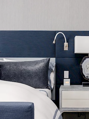 A modern bedroom with a blue headboard, white and blue bedding, a bedside table with a lamp, and a wall-mounted reading light.
