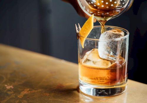 A cocktail being poured into a glass with ice and garnished with a citrus peel on a toothpick, on a bar counter.