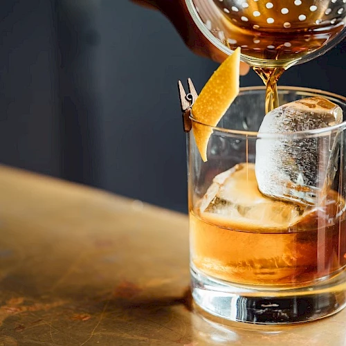 A cocktail being poured into a glass with ice and garnished with a citrus peel on a toothpick, on a bar counter.