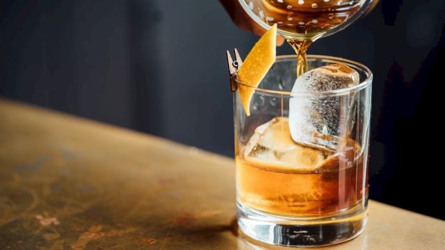 A cocktail being poured into a glass with ice and garnished with a citrus peel on a toothpick, on a bar counter.