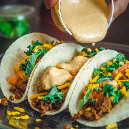Three tacos with meat, cheese, lettuce, and sauce being poured on top, served on a glass plate.