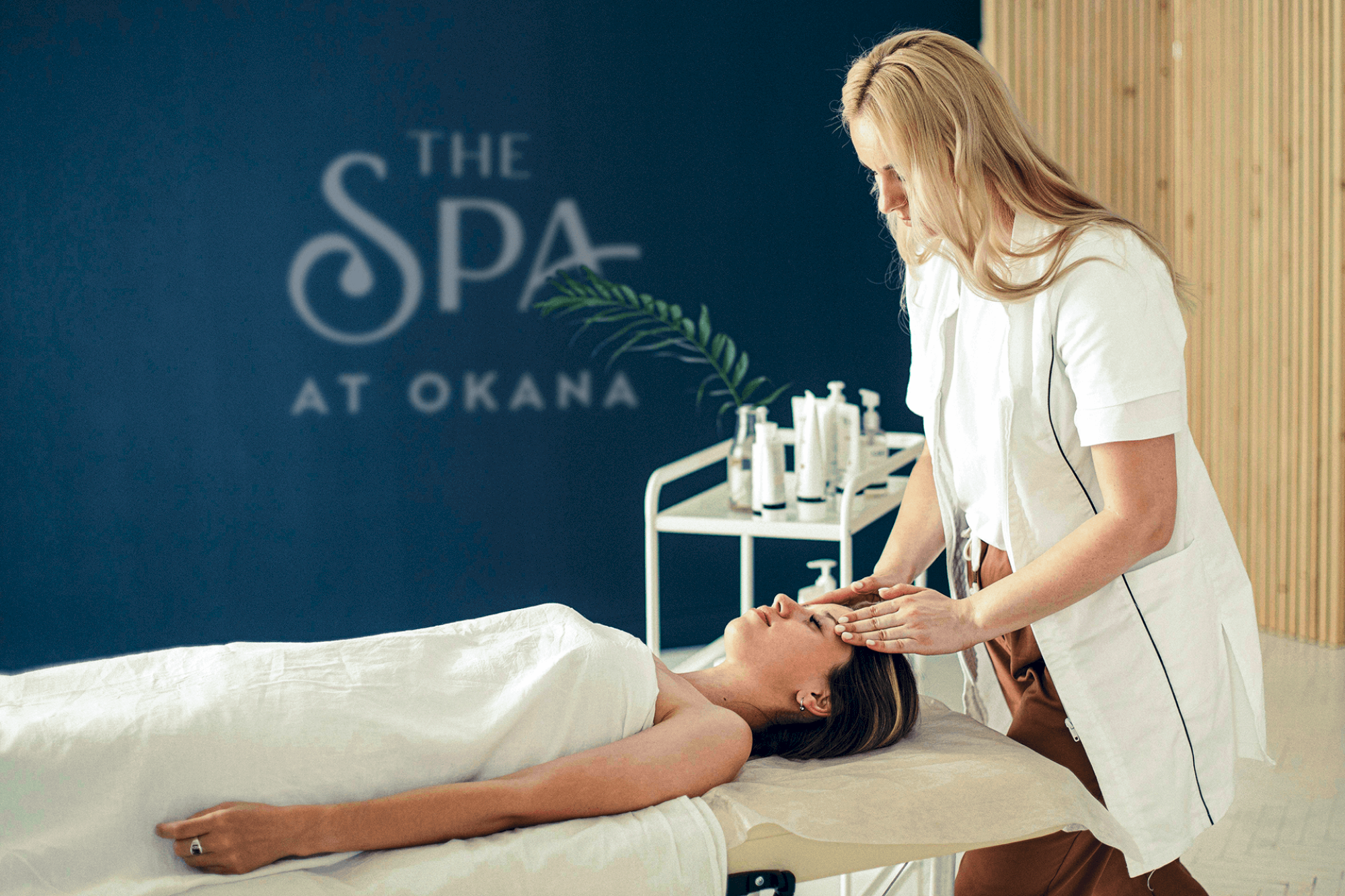 A spa setting with a person receiving a facial massage while lying on a table covered with a white sheet.