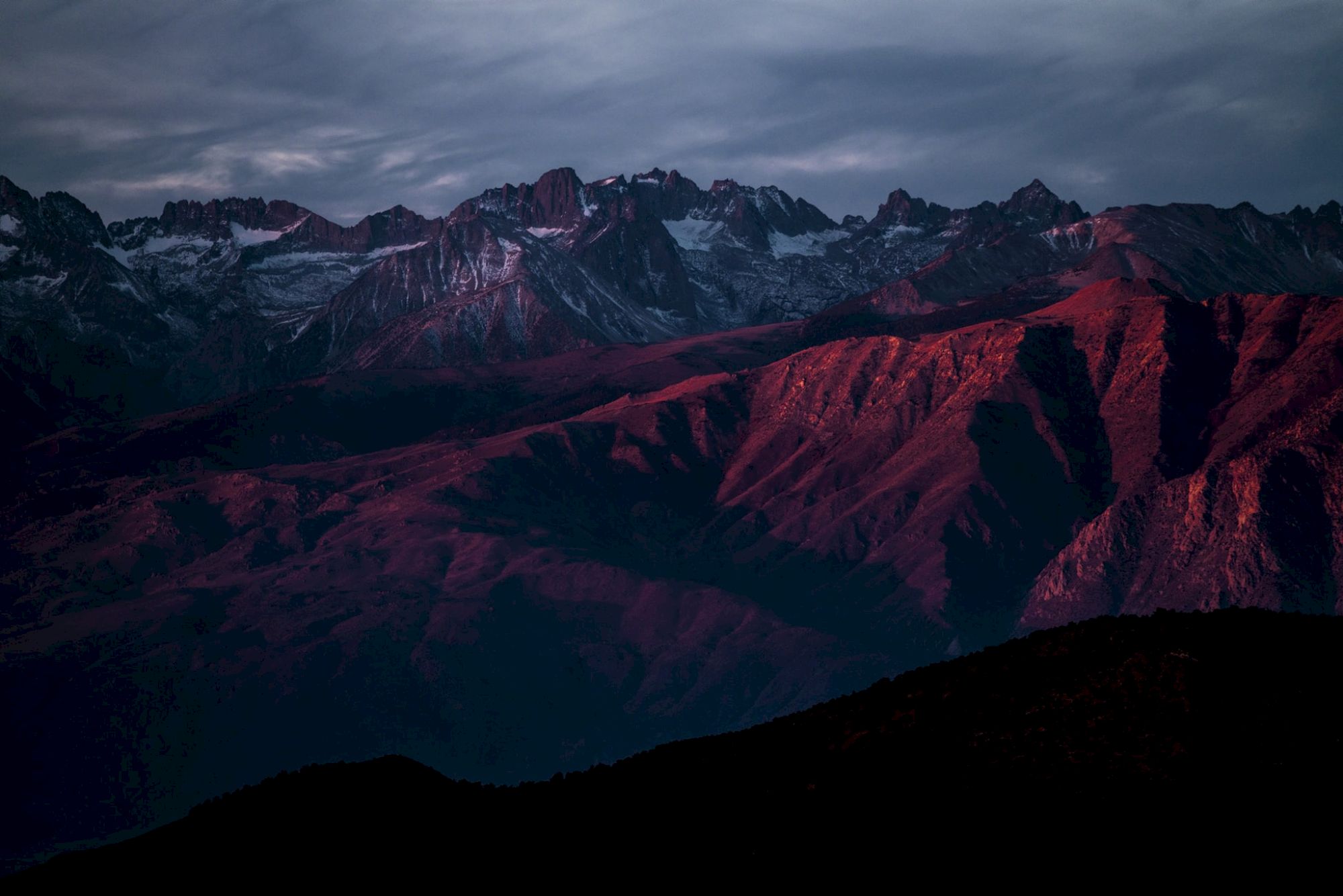 A mountain range is bathed in soft, reddish light under a dramatic, cloudy sky.
