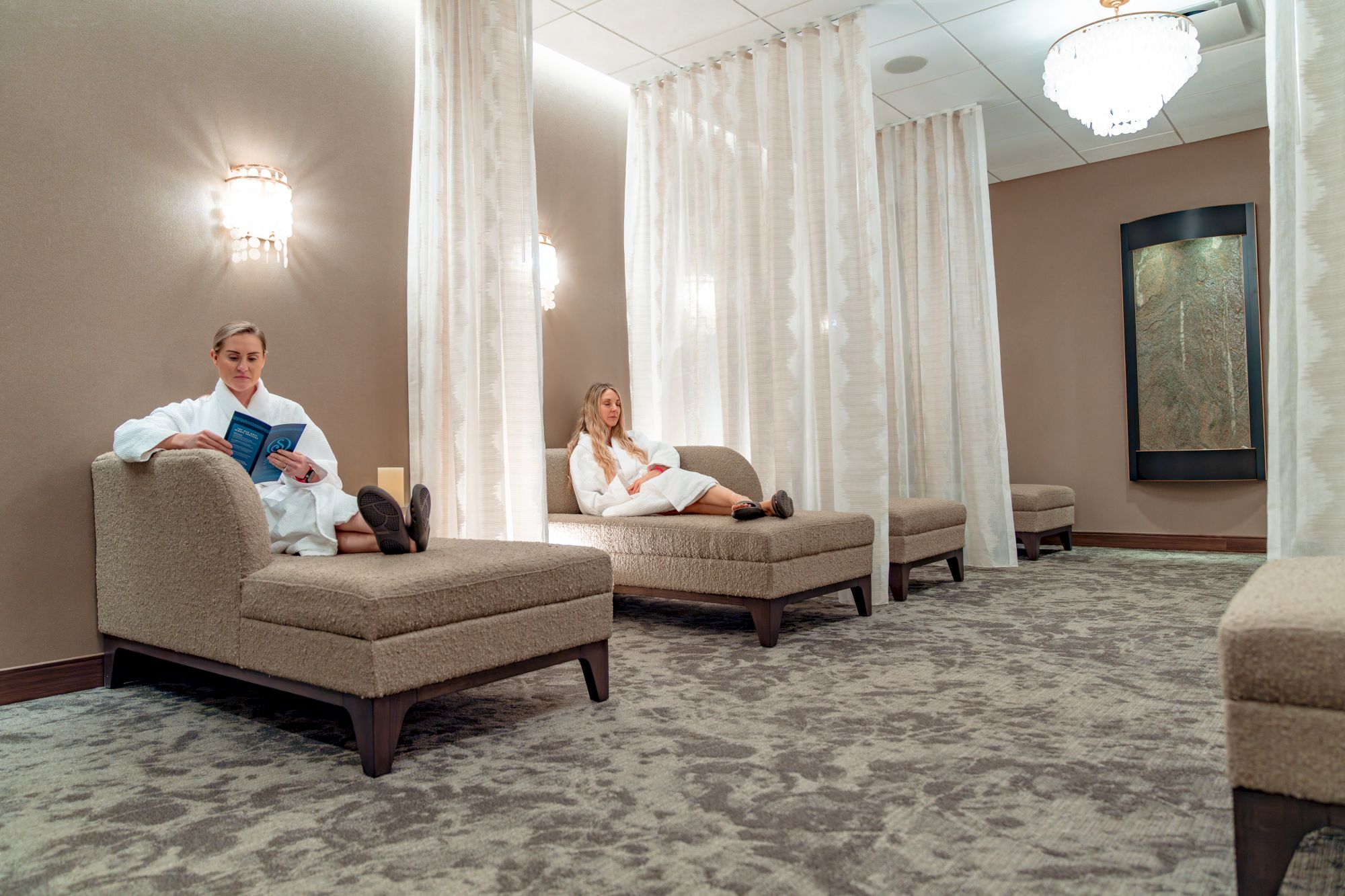 Two individuals relax on chaise lounges in a serene room with soft lighting and curtains.