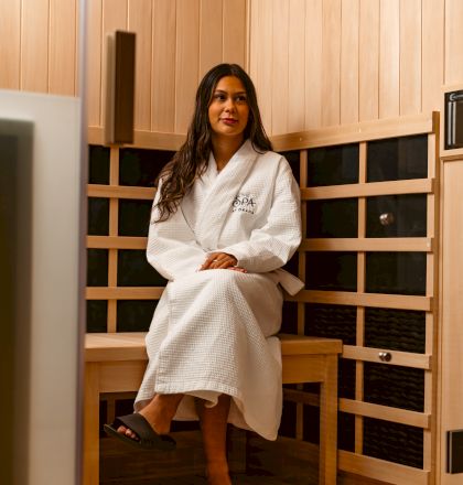 A person in a white robe and slippers sits in a wooden sauna, appearing relaxed.