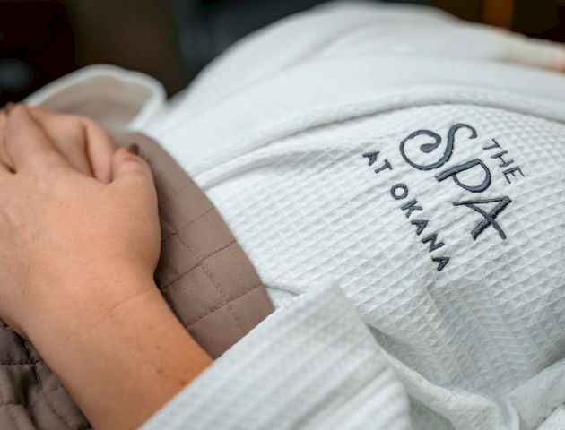 A person is wearing a white robe with "The Spa at Okana" embroidered on it, hands resting on a brown blanket.