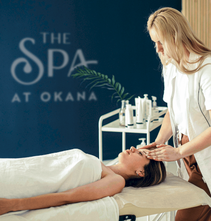 A person is receiving a facial massage at a spa, with "The Spa at Okana" visible in the background on the wall.