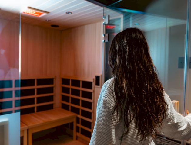 A person in a white robe opens the door to a wooden infrared sauna, revealing benches inside the warmly lit cabin.