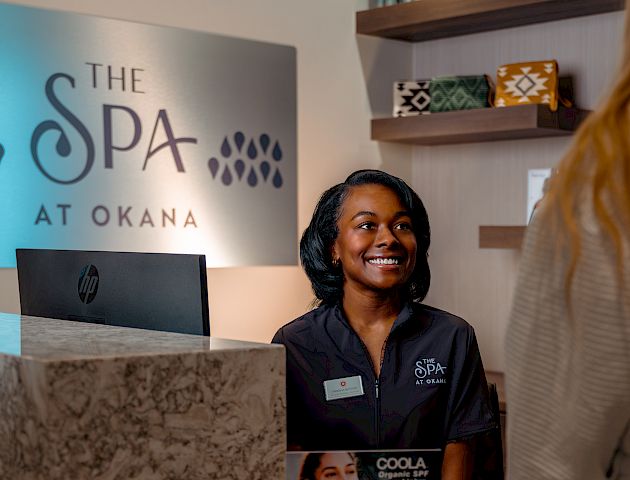 A receptionist smiles behind a counter at "The Spa at Okana." Skincare products are displayed nearby and on shelves in the background.