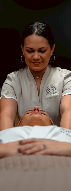 A person is receiving a massage from a practitioner wearing a "The Spa at Okana" uniform in a serene setting.