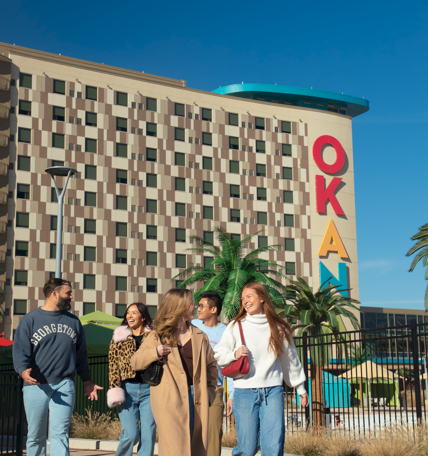 Group of friends walking outside a colorful hotel with a tall &ldquo;OKA&rdquo; sign; sunny day, palm trees, blue sky, casual outfits.