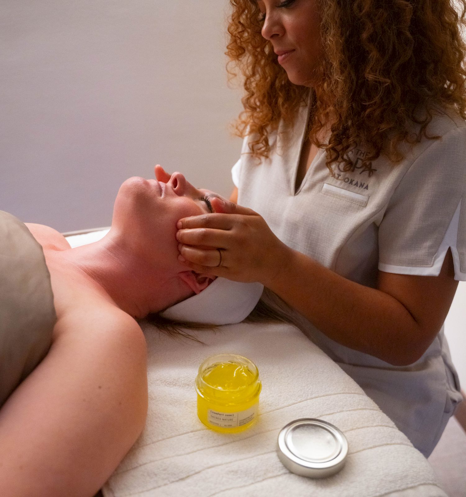A person receiving a facial treatment from an aesthetician, with a jar of cream nearby on a treatment table.