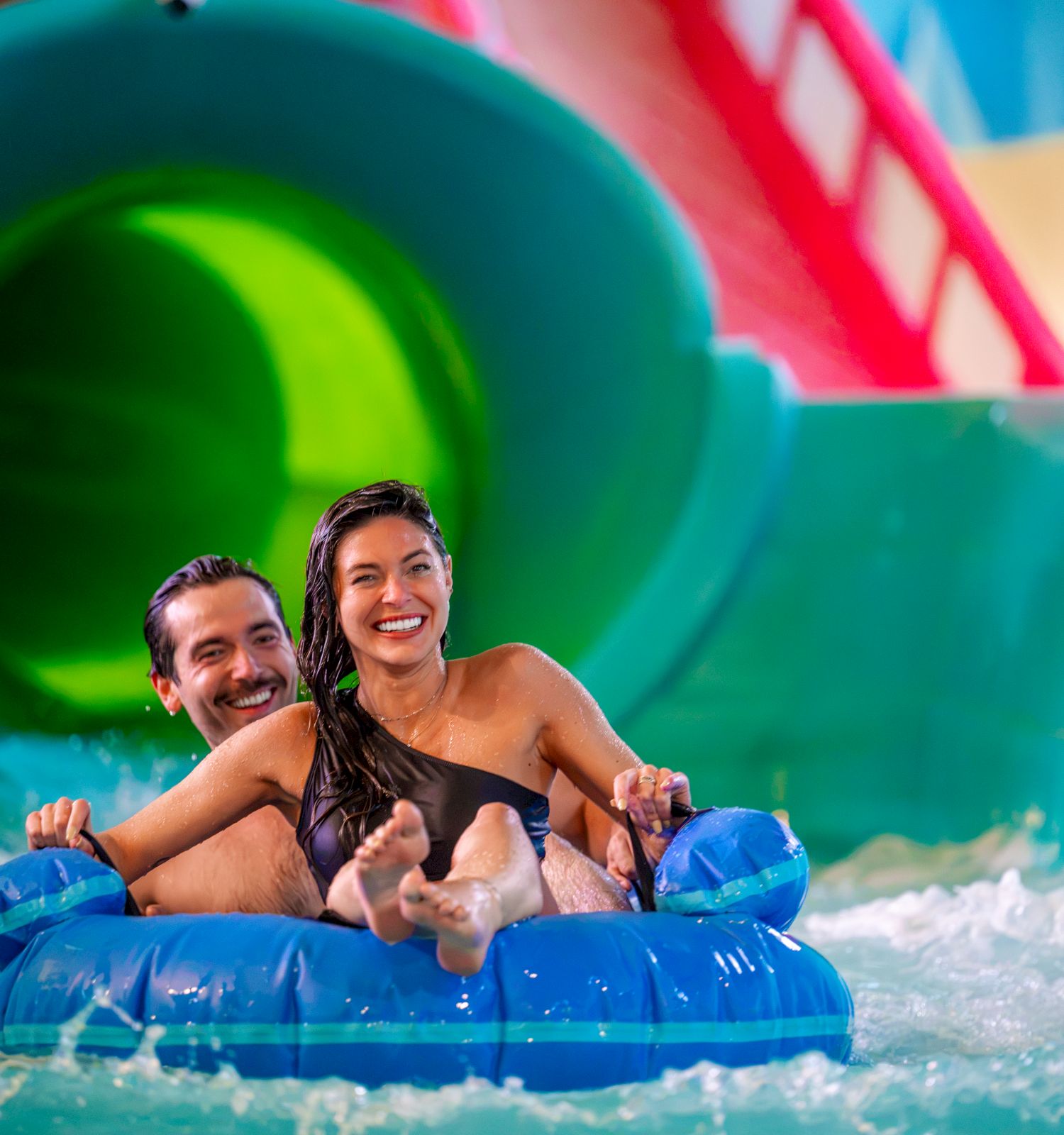 A couple smiles while riding an inflatable inner tube in a colorful water-park pool area.
