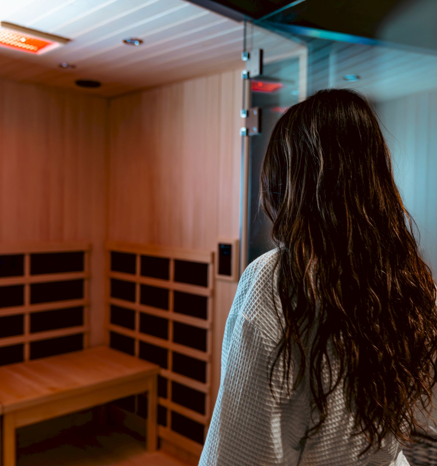 A person in a bathrobe stands at the entrance of a wooden sauna, ready to enter, with infrared panels and a bench inside.