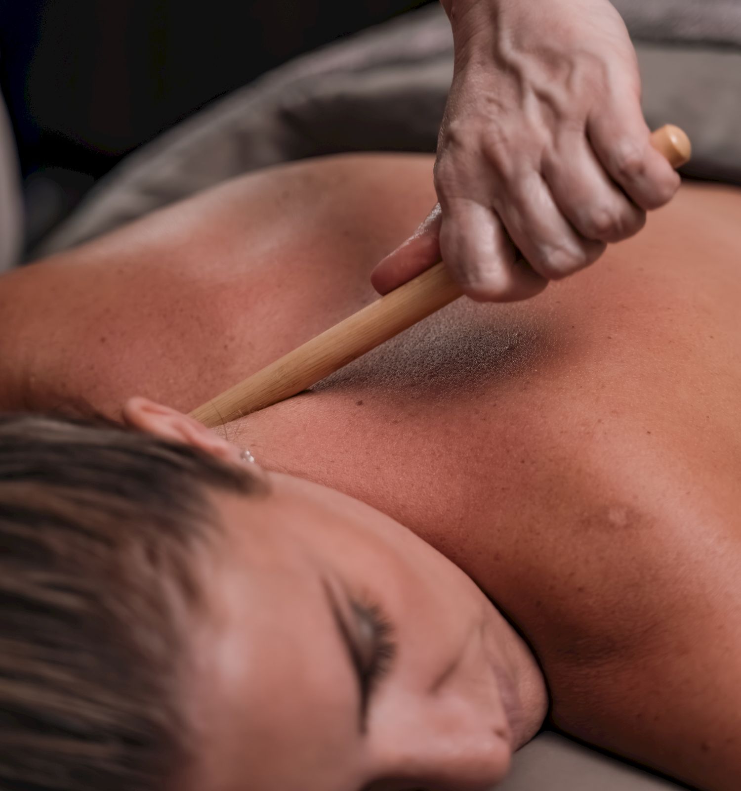 A person is receiving a massage, with a wooden tool or wand pressed along the back and shoulder area, in a dim, private setting, ending the sentence.