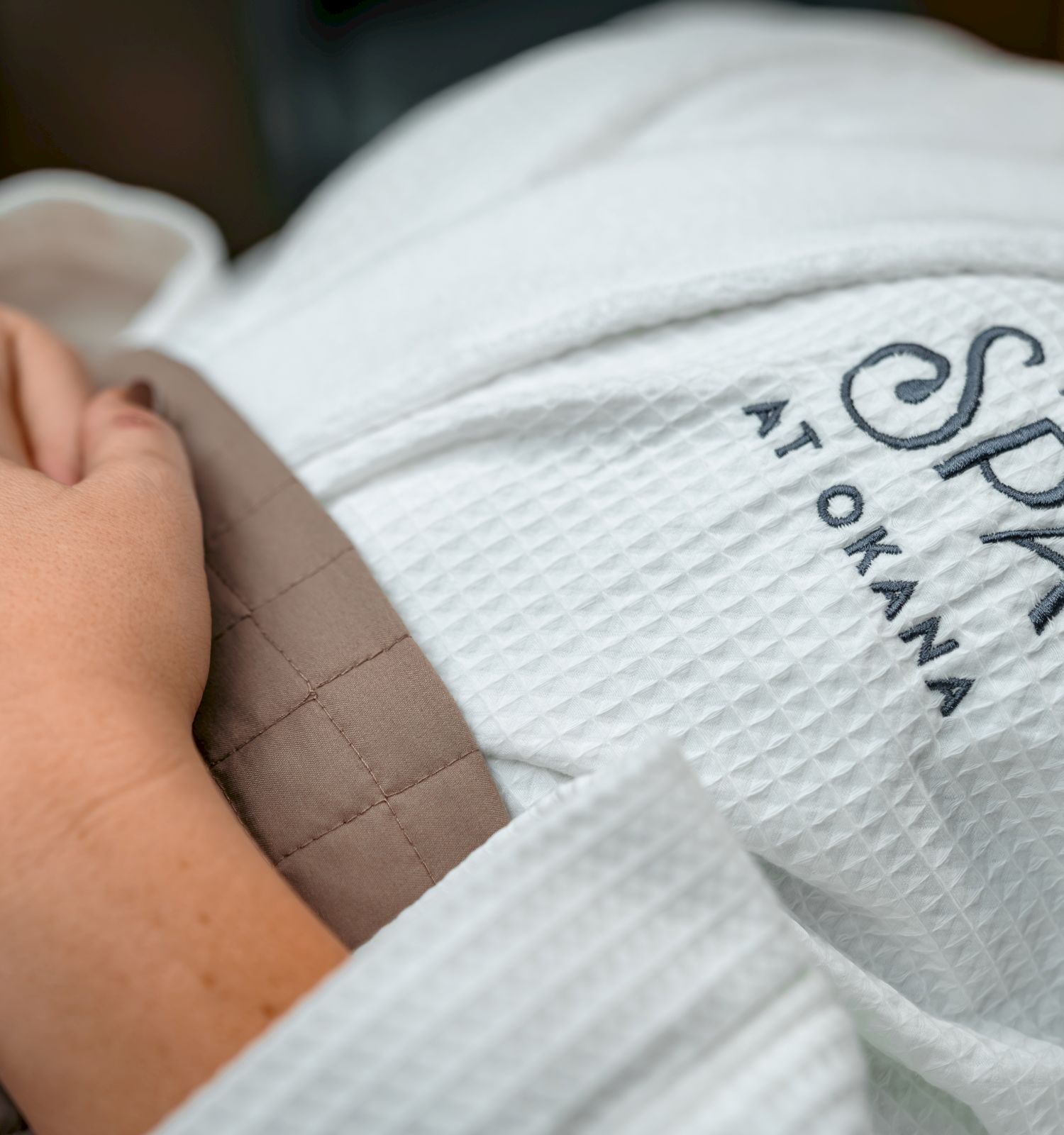 A person wearing a white robe with "The Spa at Okana" embroidered on it is resting, hands folded on their chest.