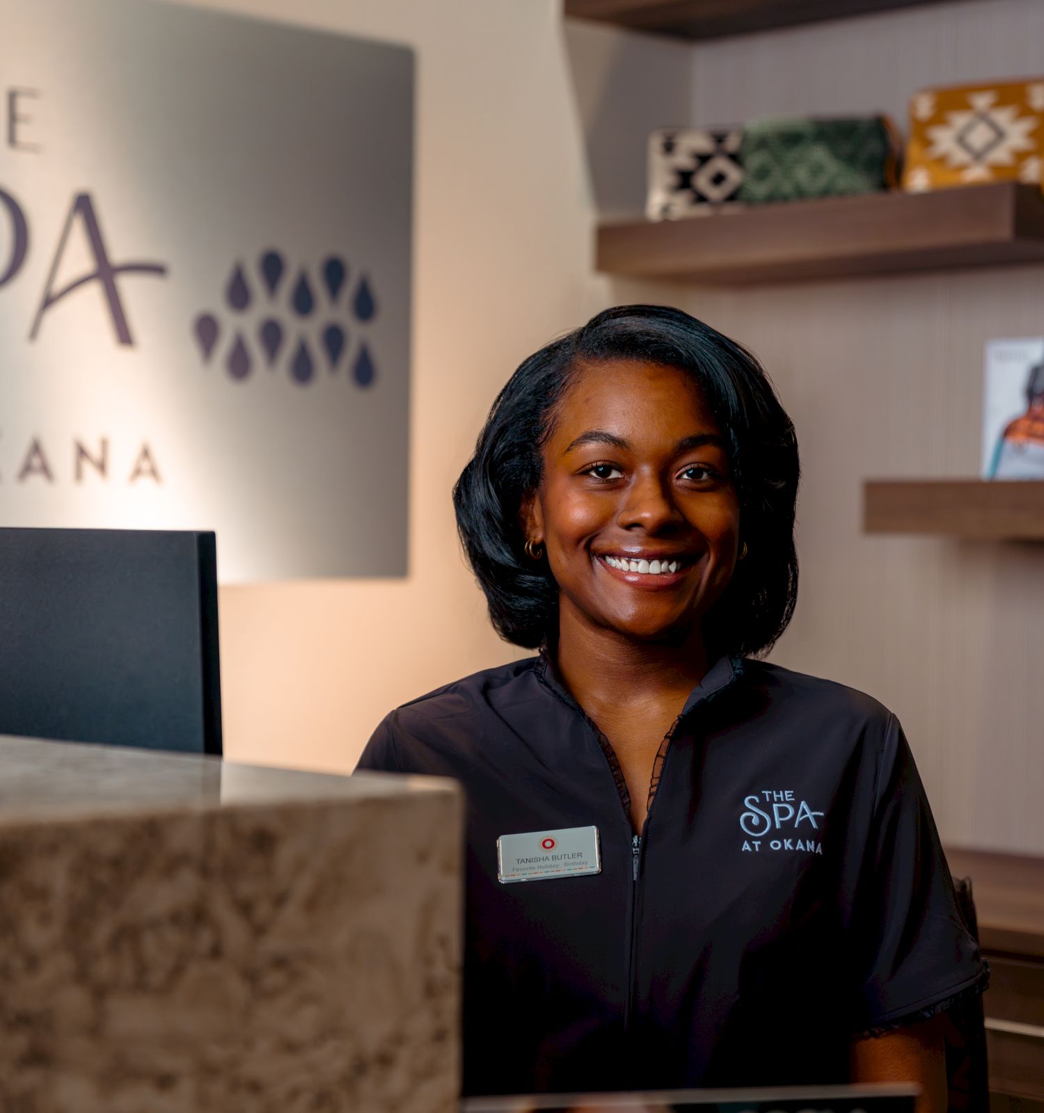 A smiling staff member at a spa reception desk with a sign reading "The Spa at Okana" in the background. Shelves display products.