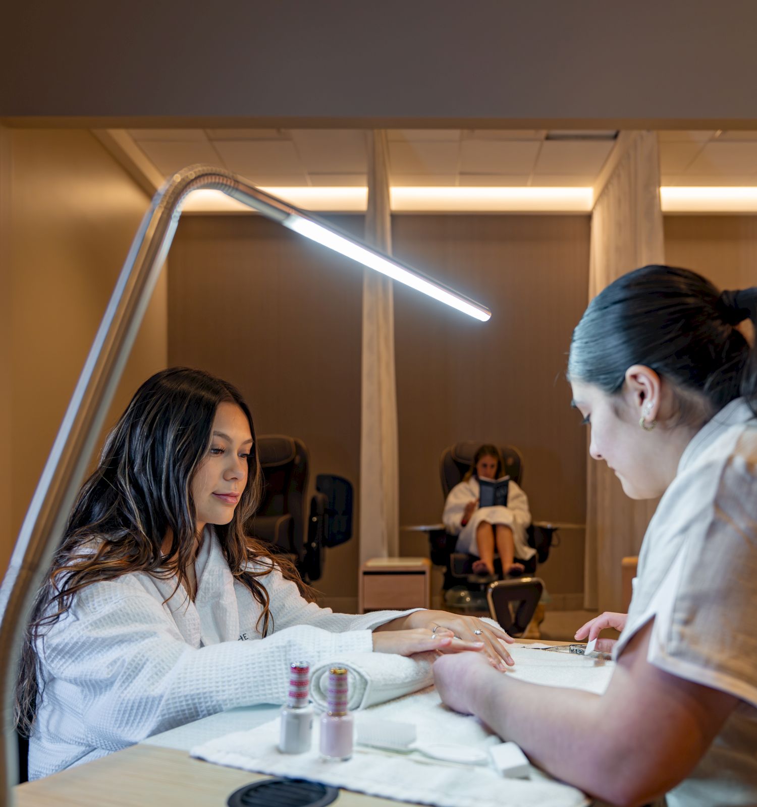 Two people at a nail salon, one getting a manicure. Another person is seated in the background under a glowing lamp.