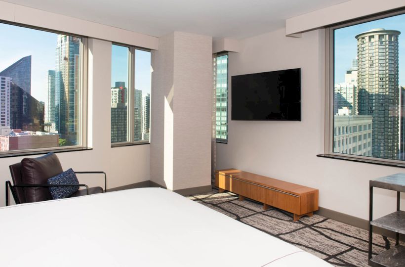 A modern hotel room features large windows with a city view, a wall-mounted TV, a bed, a chair, and a small table.