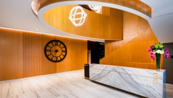 Modern lobby with a sleek marble reception desk, spiral stairs, wall clock, and decorative lights, enhanced by a flower vase.