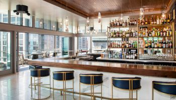 The image shows a modern bar with stylish seating, a well-stocked shelf, and large windows offering a city view.